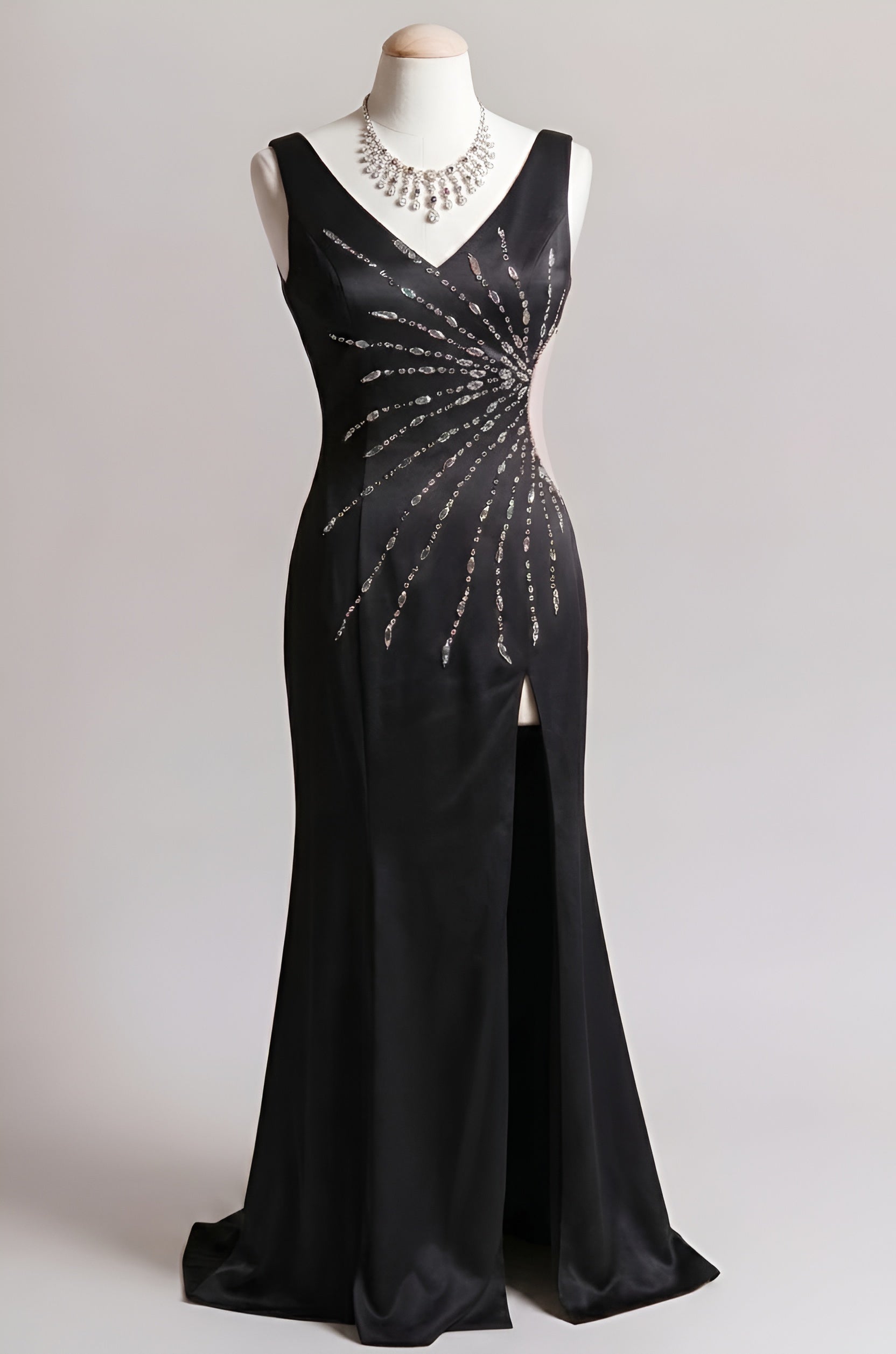 A floor-length black evening gown displayed on a mannequin against a light gray studio background. The sleeveless V-neck dress features a fitted silhouette with a high slit and a decorative radial line of sparkling embellishments across the bodice and skirt.